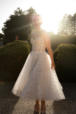 A woman wearing a strapless white lace dress with a fitted bodice and full skirt, standing outdoors in front of bright sunset light with trees and hedges in the background.