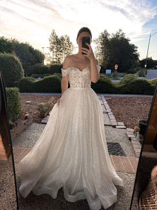 A woman takes a mirror selfie wearing an off-the-shoulder A-line wedding dress with a lace sweetheart bodice and a full, sparkly tulle skirt, set against an outdoor garden background at sunset.