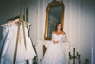 A woman wearing a white off-the-shoulder lace wedding gown stands in a decorated room with white walls, a large gold-framed mirror, and a dress rack with multiple white gowns to her left.