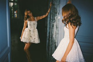 A woman wearing a white sleeveless lace dress stands in a dim interior doorway while looking at her reflection, which shows the full length of the dress. The photo has a vintage, Polaroid-style border and cool blue-toned lighting.