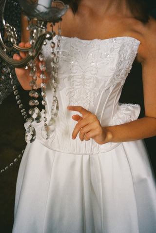 A white strapless wedding dress with a heavily embellished lace and bead corset bodice, worn by a model holding a silver chandelier.